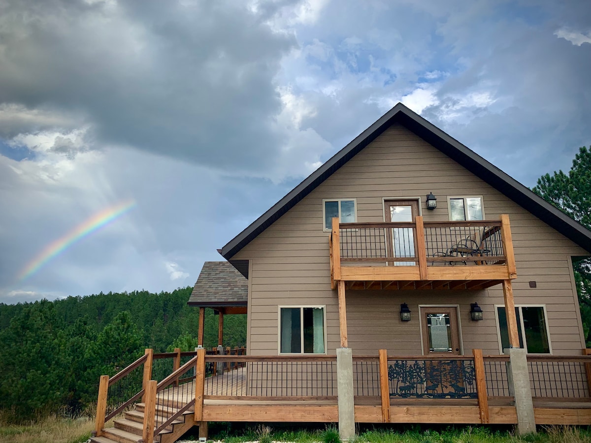 A newly-built cabin features a welcoming front porch with wooden steps leading to an elevated deck. The exterior showcases neutral tones and large windows, with a backdrop of lush forest. A faint rainbow arcs across the sky, enhancing the tranquil setting.