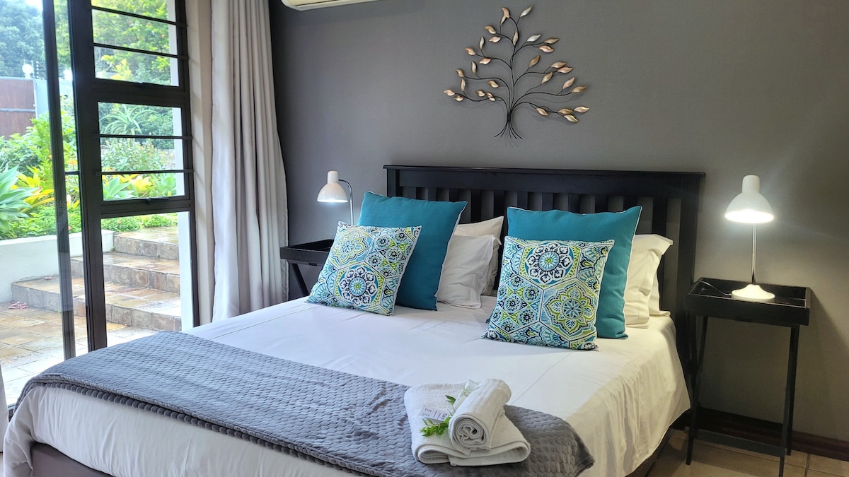 A comfortable double bed is dressed in crisp white linens, complemented by vibrant blue and patterned throw pillows. A decorative wall piece adds charm, while natural light filters through large windows, offering views of the garden. Two bedside lamps provide soft illumination.