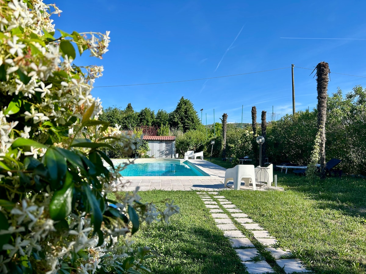 A serene garden scene captures a sparkling pool surrounded by vibrant greenery. White lounge chairs and a small table are positioned nearby. The area is framed by blooming flowers and mature trees, with a clear blue sky lending a sense of openness.