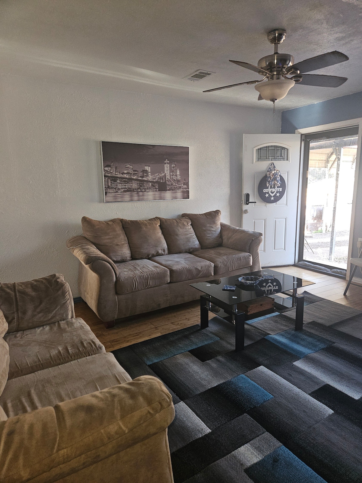 A comfortable living area features a plush sofa and loveseat positioned around a low coffee table. A wall art piece adds interest above the sofa. Natural light enters through a door that leads to an outdoor space, while a ceiling fan is installed for air circulation.