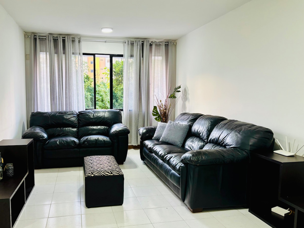 A bright living area features two black leather sofas and a small storage ottoman. Natural light enters through a large window, framed by sheer curtains, creating a soft glow. A potted plant adds a touch of greenery, complemented by a dark cabinet against the wall.