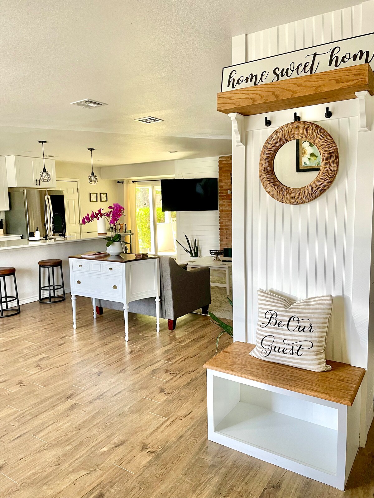 An inviting entryway features a bench with a sign that reads 'home sweet home.' A round mirror is mounted above the bench, reflecting light. To the left, a kitchen area is visible with bar stools at the counter, and a cozy seating area can be seen in the background.