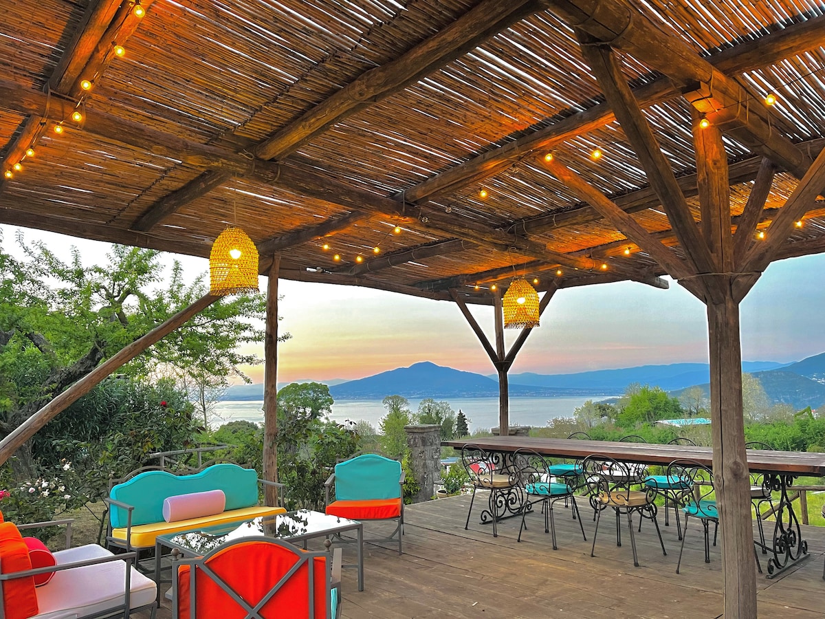 A shaded outdoor seating area features colorful chairs and a wooden table, illuminated by soft hanging lights. The backdrop showcases a tranquil view of the sea and distant mountains, reflecting the soft colors of dusk.