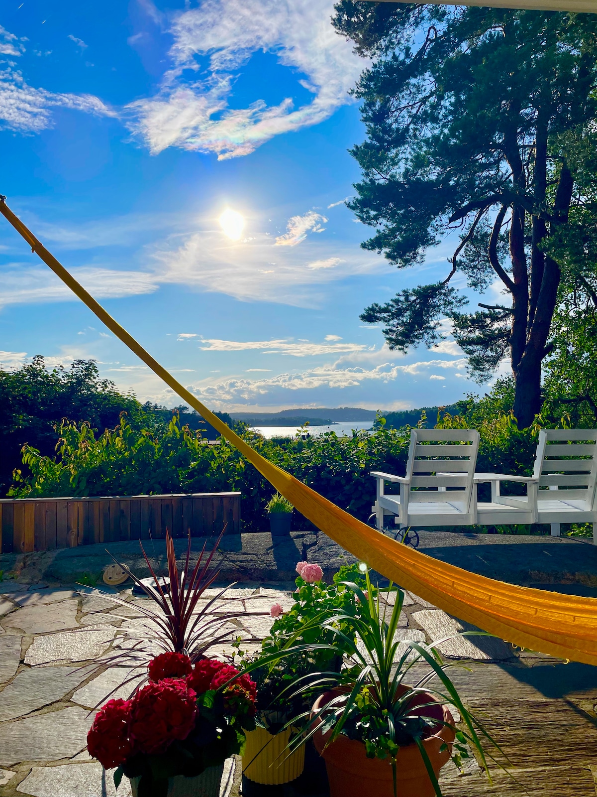 A spacious outdoor area features a hammock strung between two trees, with colorful flowers in pots nearby. White chairs are positioned to offer a view of the landscape, including the water and sky, under bright sunlight and scattered clouds.