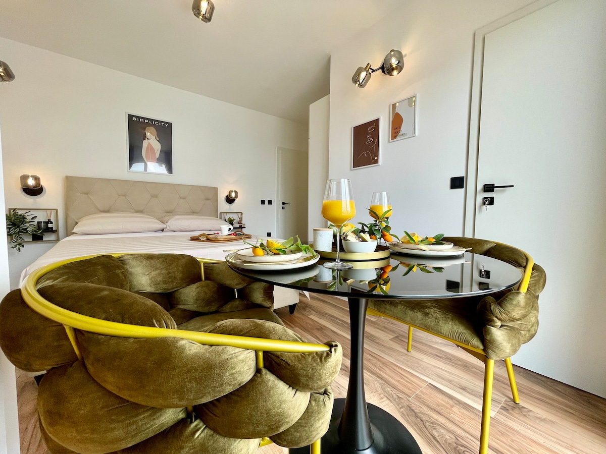 A small dining area is set with two plush chairs surrounding a black table. A fresh breakfast is displayed, accompanied by two glasses of orange juice. In the background, a soft, light-coloured bed is visible, complementing the modern decor.