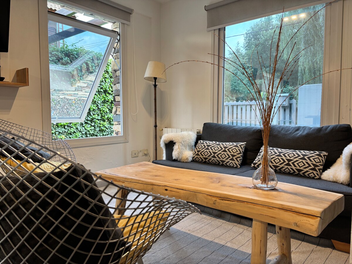 A comfortable living area features a dark sofa adorned with decorative pillows, complemented by a wooden coffee table. A large window invites natural light, providing views of the greenery outside. A floor lamp stands beside the seating, adding a cozy element to the space.
