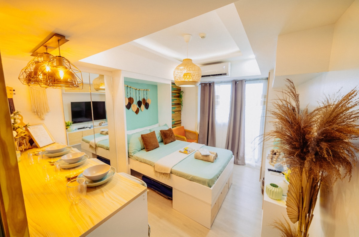 An open living space features a comfortable bed adorned with light aqua bedding and decorative pillows. A kitchen counter with dining ware is visible, alongside a seating area. Natural light fills the room through large windows, with soft curtains enhancing the ambiance.