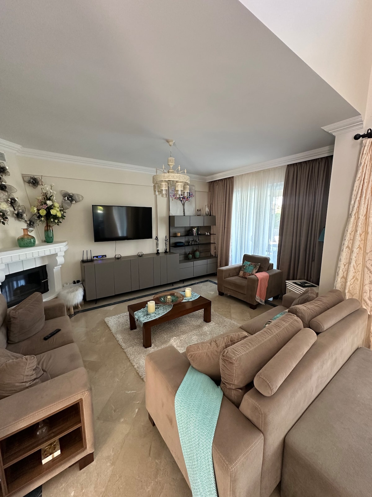 A cozy living area is highlighted by plush seating and a wooden coffee table. A modern television is mounted on the wall above a contemporary console. Soft curtains frame a window, allowing natural light to brighten the room, which is adorned with decorative accents.
