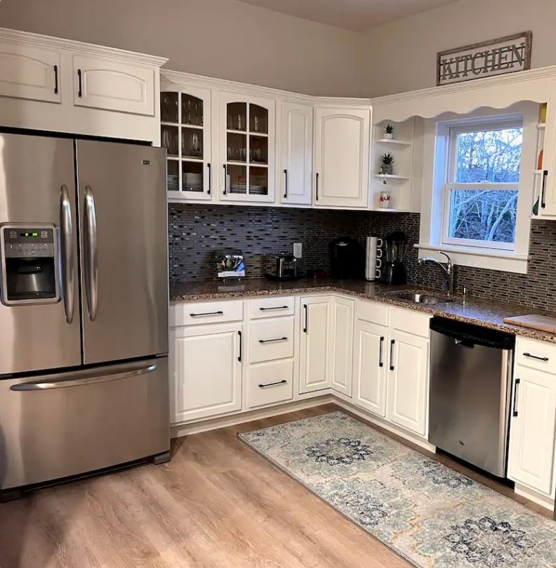 A modern kitchen is displayed, featuring white cabinetry and stainless steel appliances. A large refrigerator and a dishwasher are positioned next to a stylish countertop. Natural light filters in through a window above the sink, complementing the patterned rug on the wooden floor.