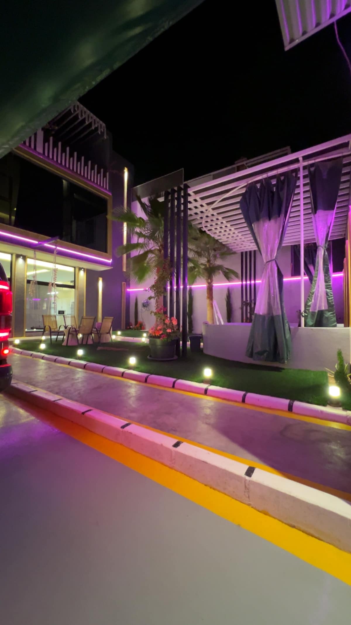 An illuminated exterior showcases modern architecture with vibrant purple lighting. A spacious terrace area features stylish seating surrounded by greenery, while curtains drape elegantly. Pathway lighting highlights the approach to the entrance, blending seamlessly with the contemporary aesthetic.