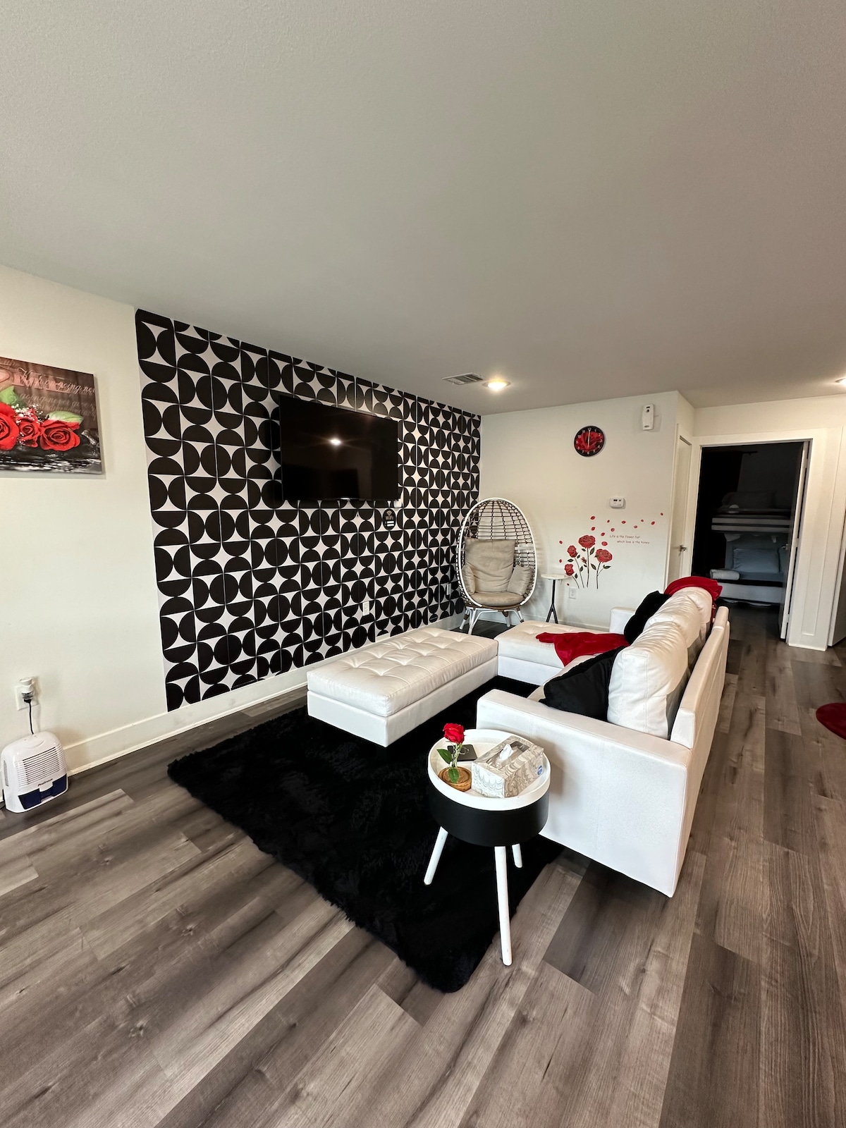 The living room showcases a striking feature wall with geometric patterns alongside a mounted TV. A white sofa is paired with a matching ottoman and a small side table adorned with roses. A hanging chair adds a unique touch, complemented by modern flooring.