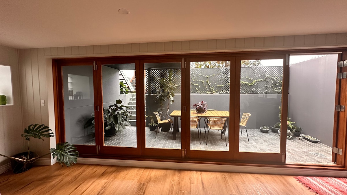 A private patio is visible through large glass doors, allowing natural light to flow into the space. The outdoor area features a wooden dining table accompanied by several chairs, surrounded by lush greenery. The flooring consists of wooden planks, enhancing the inviting atmosphere.
