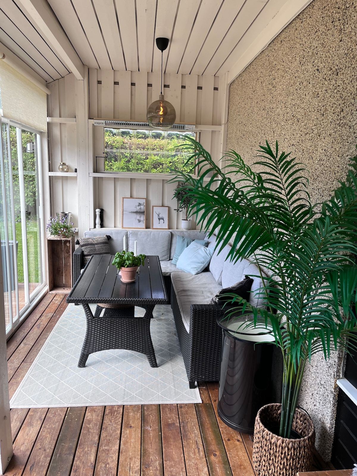 A charming sunroom features a comfortable seating area with a dark wicker sofa and a coffee table. A potted plant adds a touch of greenery, while light streams in through large glass doors, creating a bright and inviting space. Wooden flooring complements the decor.