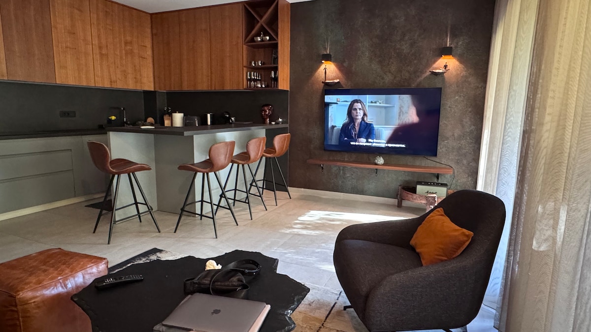 The living area features a modern kitchen with wooden cabinetry and high stools at the bar. A comfortable chair with an orange accent pillow stands next to a dark coffee table. A flat-screen TV is mounted on the wall, enhancing the overall design.