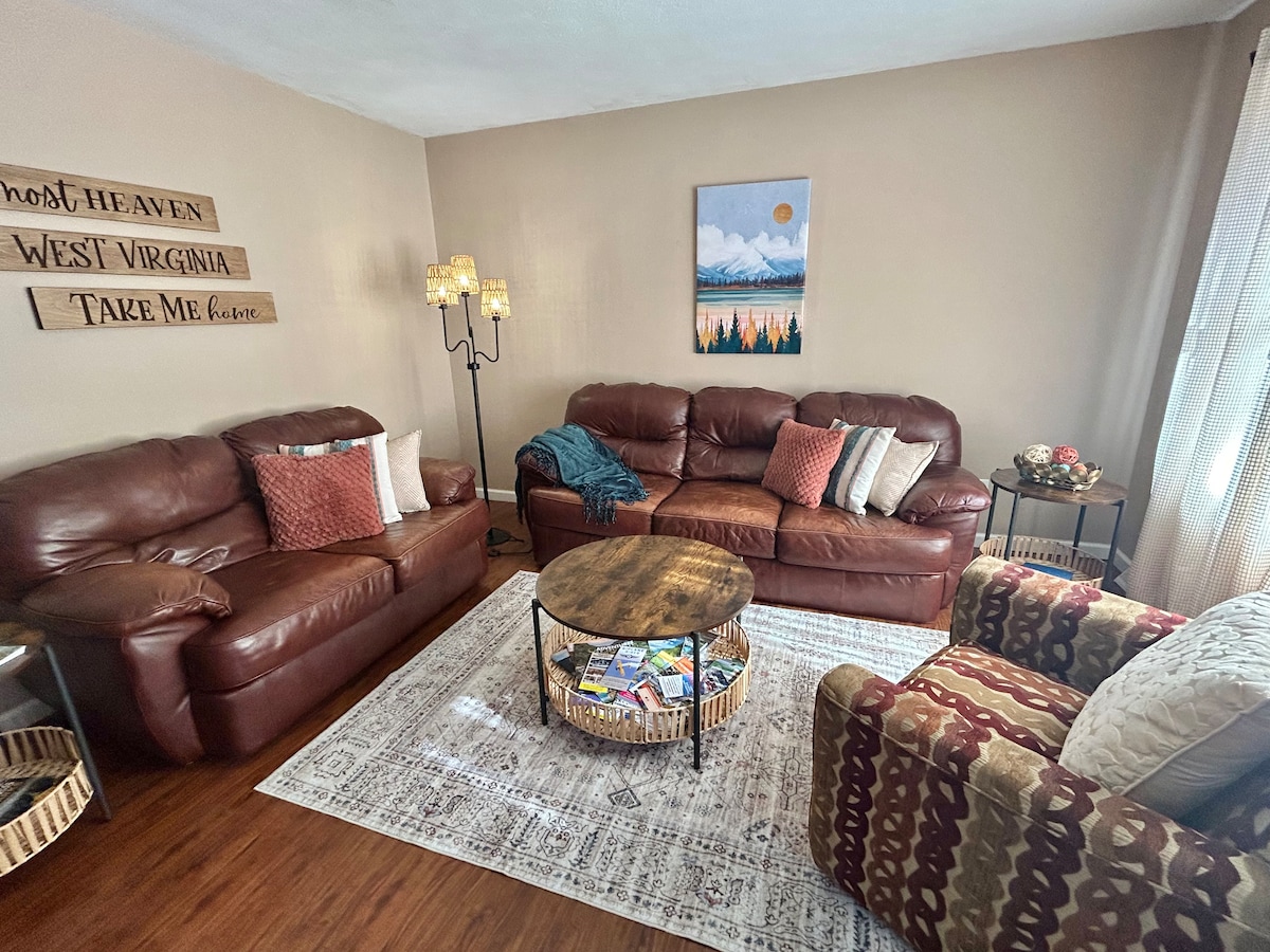 A cozy living room showcases two leather sofas and a patterned accent chair, accompanied by a round coffee table adorned with magazines. Light streams in through a window, illuminating the warm decor featuring mountain artwork and decorative wall signs.