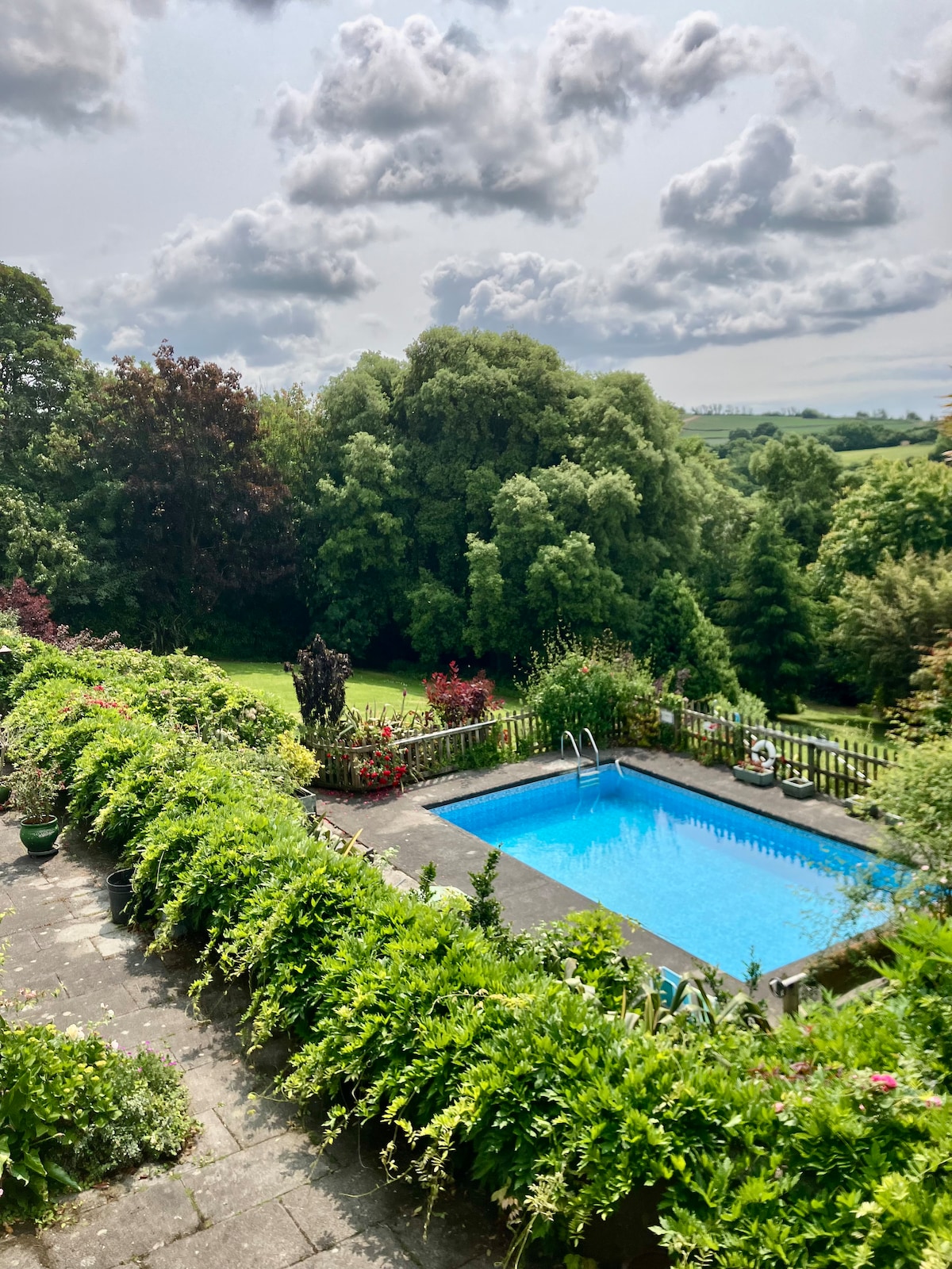 A swimming pool is situated in the garden area, surrounded by lush greenery and colorful blooming flowers. Views of trees and rolling hills are visible in the background under a partly cloudy sky. The patio area features stone tiles and is outlined with planted greenery.