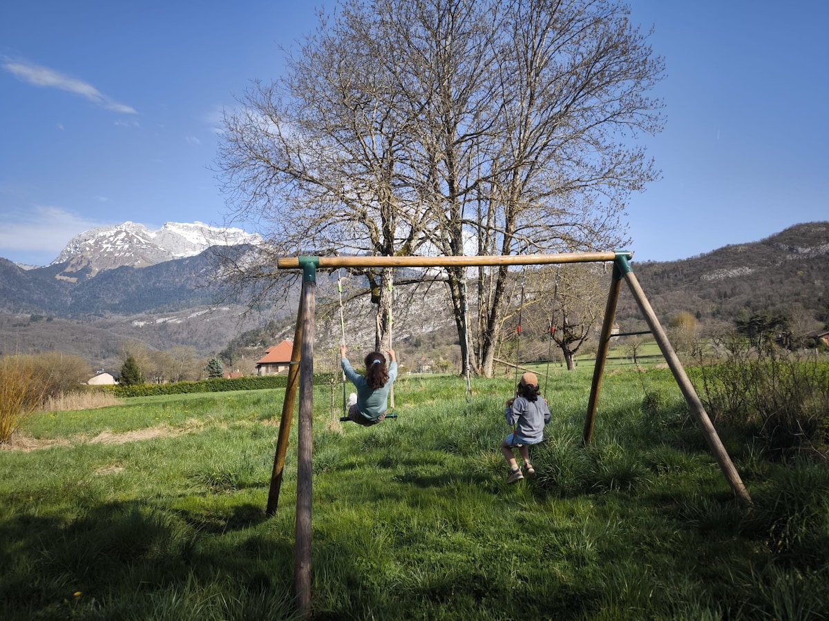 A swing set is situated in a grassy area, surrounded by trees and mountains. One child is swinging while another child stands nearby, both enjoying the outdoor space. The clear blue sky enhances the natural landscape.
