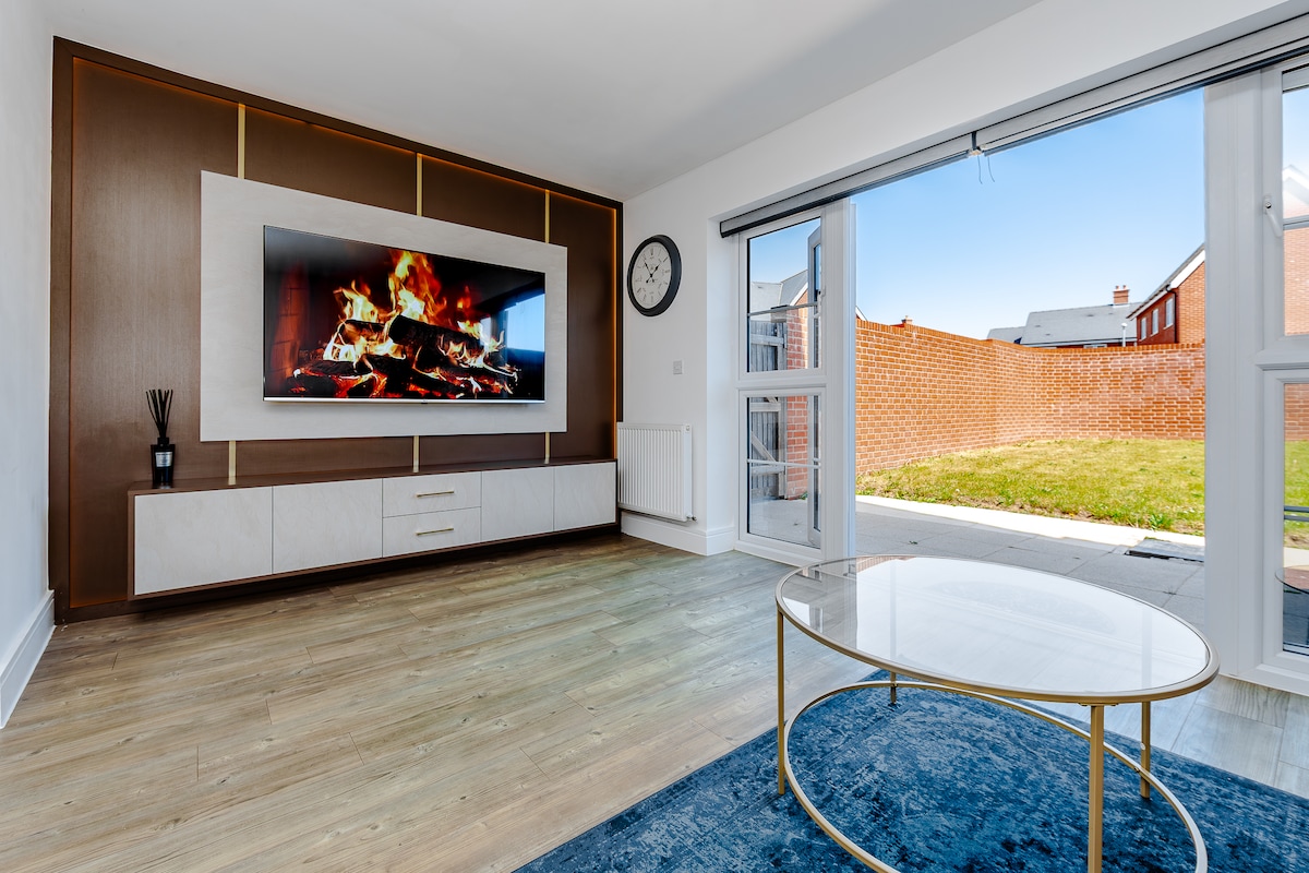A bright living space features a modern wall unit with a large, wall-mounted TV displaying a fireplace. A circular coffee table is positioned on a blue area rug, and sliding glass doors open to a private outdoor area with green grass.