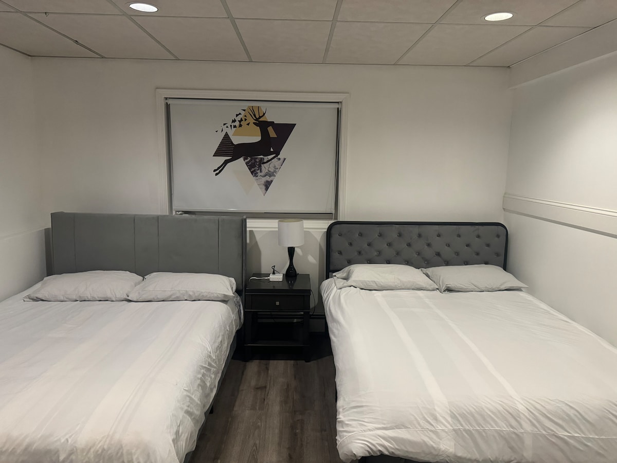 A room features two spacious beds, one with a tufted headboard. A nightstand with a lamp is positioned between the beds. A colorful wall art piece is displayed above the window, brightening the minimalist white walls and dark flooring.