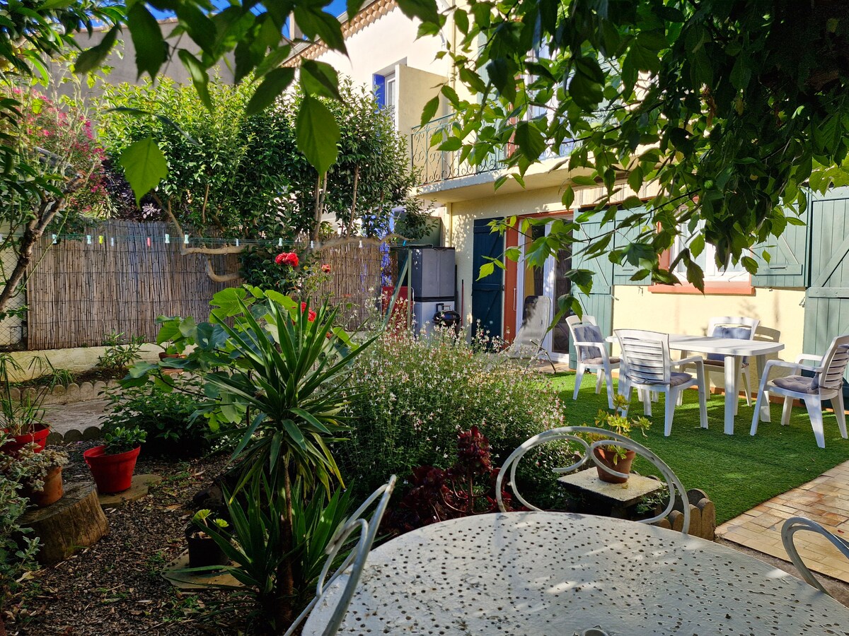 A serene garden setting is presented, featuring a round table with intricate patterns and white chairs. Lush greenery surrounds the area, complemented by colorful flowers and potted plants. The garden is enclosed by a natural fence, enhancing the tranquil outdoor space.