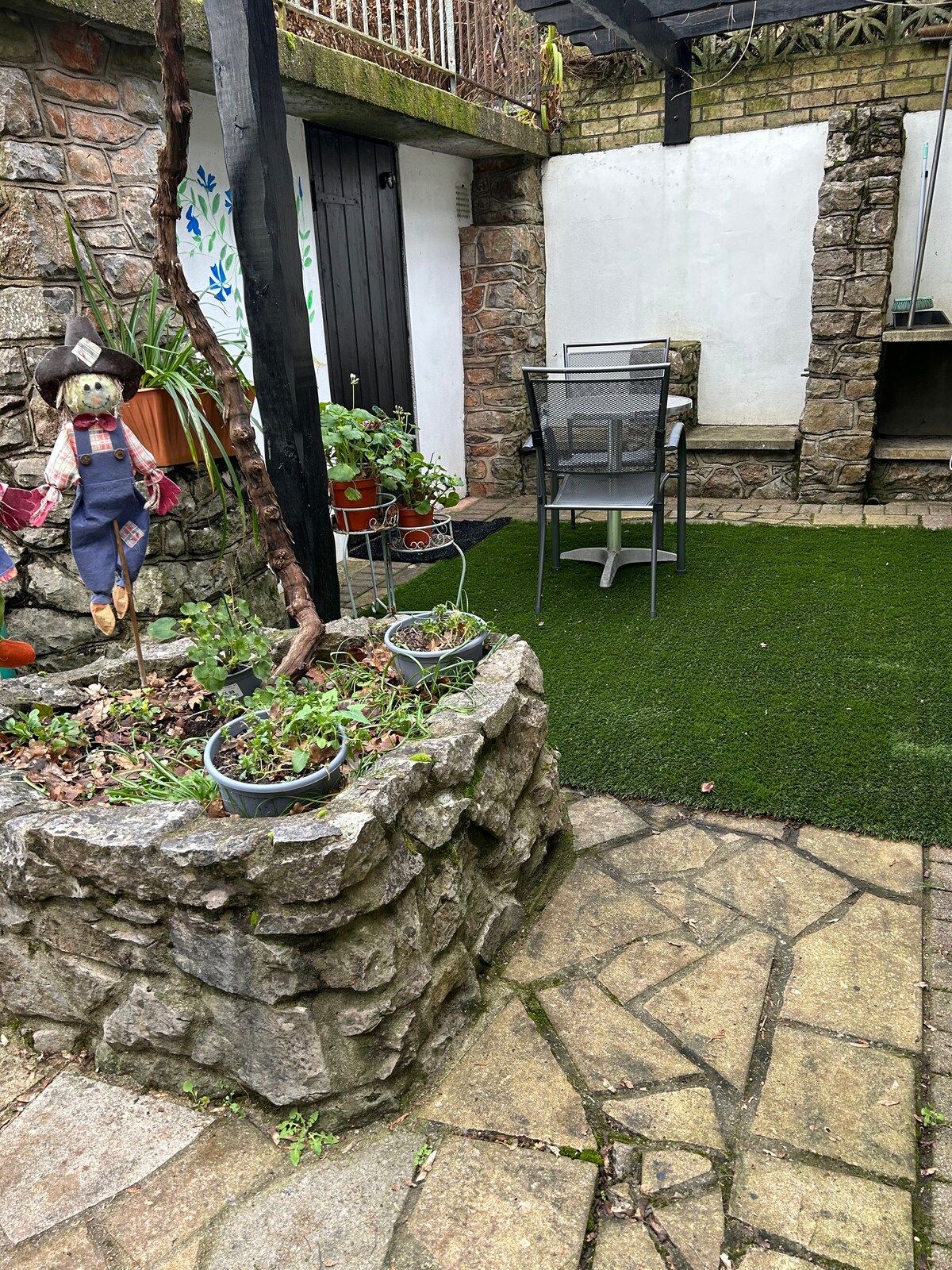 A small outdoor area is visible, featuring stone pathway tiles leading towards a grassy patch. Potted plants adorn a stone planter, while a dining set with a glass table and chairs is positioned in the corner, creating a cozy space for relaxation.