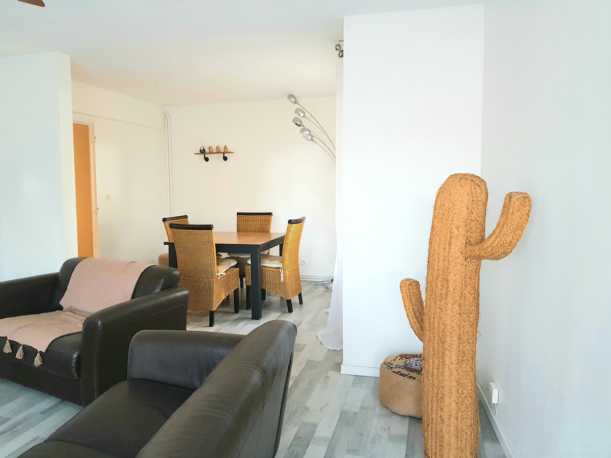 A spacious living area features a black leather couch and a woven straw cactus decoration. A dining table with four chairs is positioned near a wall, illuminated by modern floor lamps. Light-colored flooring complements the bright, open atmosphere of the room.