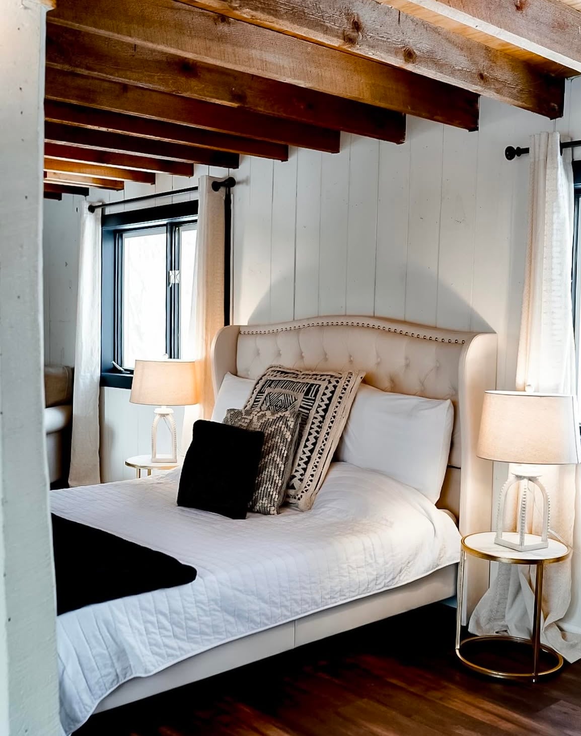 A cozy sleeping area features a queen bed with a tufted headboard and layered bedding. Each side is complemented by a circular table with a lamp. Natural light enters through two windows, framed by soft, light-colored curtains, while exposed wooden beams enhance the rustic charm.