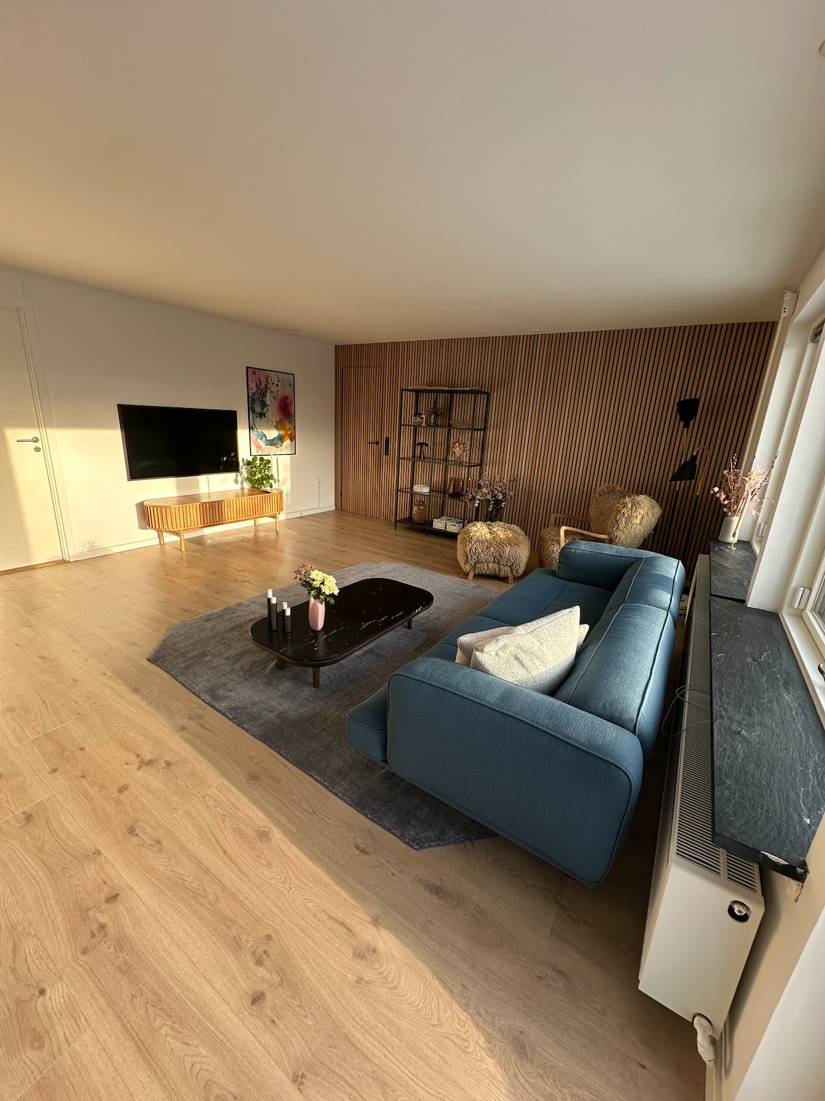 A modern living room is presented with light wood flooring. A comfortable blue sofa is positioned in front of a wall-mounted television. Decorative shelving is seen against a wood-paneled wall, while a soft area rug contrasts the flooring. Natural light enters through large windows.