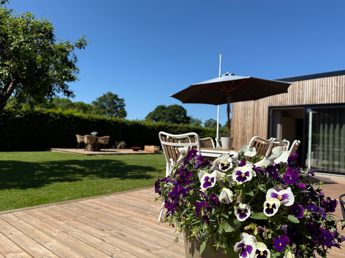 A wooden deck is adorned with a planter filled with vibrant purple and white pansies. In the background, a spacious lawn features a patio area with seating under a large umbrella, surrounded by greenery and clear blue skies.