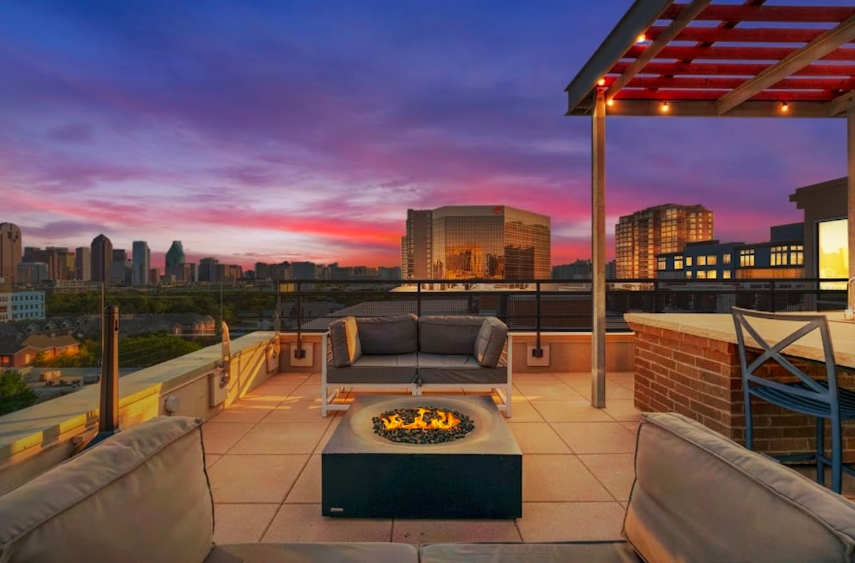 A spacious rooftop terrace showcases a comfortable seating area with contemporary sofas arranged around a fire pit. The city skyline serves as a backdrop, complemented by a vibrant sunset filled with hues of purple and orange. String lights provide a warm glow overhead.