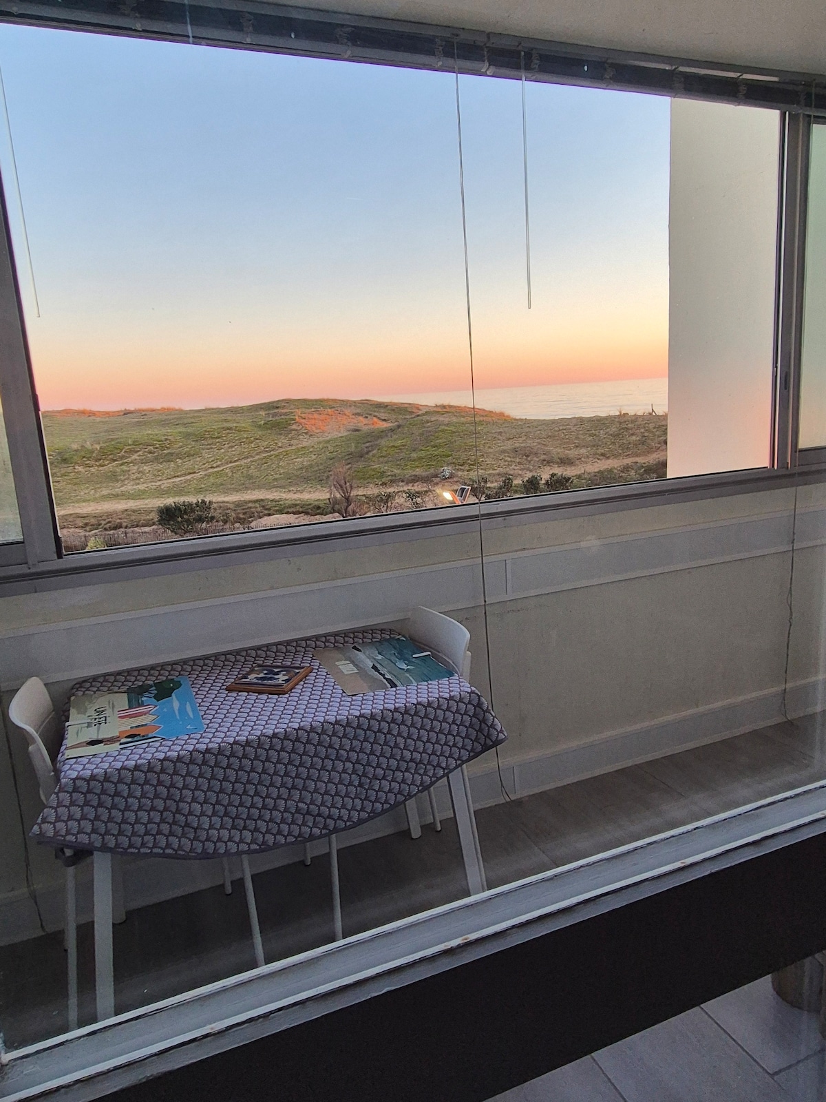 A view from the Loggia captures a serene sunset over lush dunes and the ocean. A small table with a patterned cover is positioned near the window, allowing for an intimate dining experience while enjoying the scenery. Natural light fills the space.