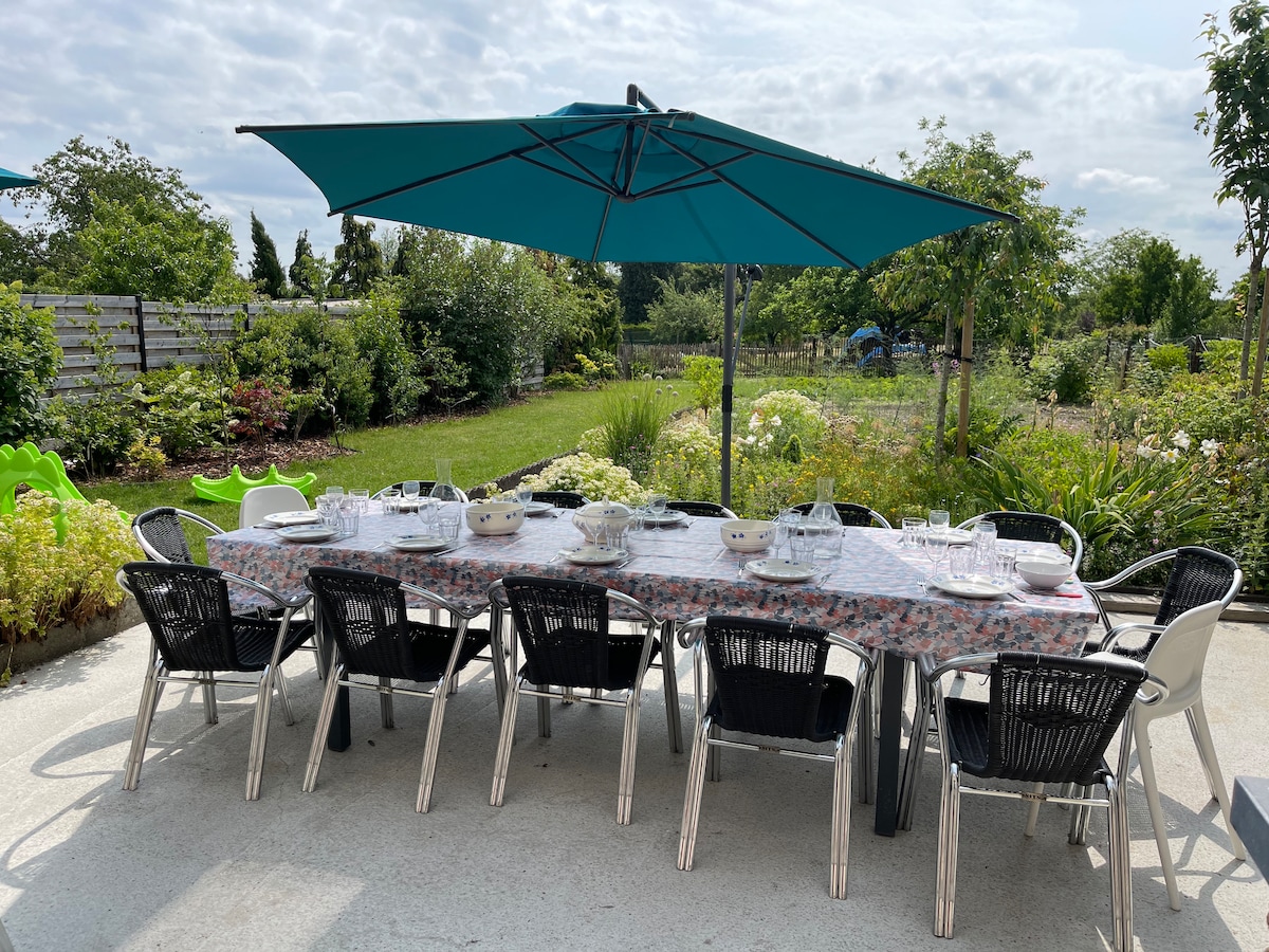 An outdoor dining area is set with a long table covered in a floral tablecloth. Multiple chairs surround the table, and a large umbrella provides shade. Lush greenery and flowering plants are visible in the background, creating a serene garden setting.