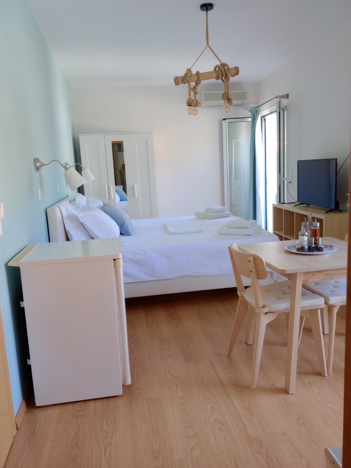 A well-lit studio features a spacious bed dressed in white linens, complemented by soft blue walls. A small dining table with two chairs is visible alongside a compact refrigerator. A wall-mounted TV is positioned near the window, allowing natural light to brighten the space.