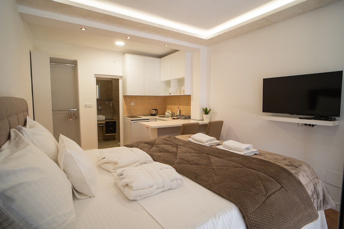 A modern studio apartment is depicted, featuring a neatly arranged sleeping area with a spacious bed dressed in neutral tones. A wall-mounted TV is visible, and the kitchenette is seen in the background, equipped with essential appliances. Soft lighting enhances the cozy environment.