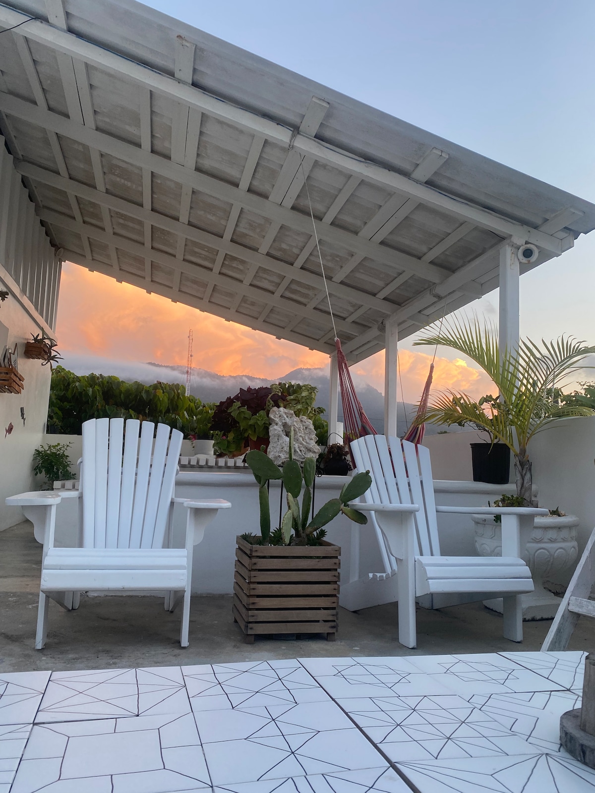 A rooftop lounge area is featured, showcasing two white Adirondack chairs positioned on a geometric tiled surface. A wooden planter with lush greenery adds a natural touch, while a colorful sunset illuminates the sky in the background.