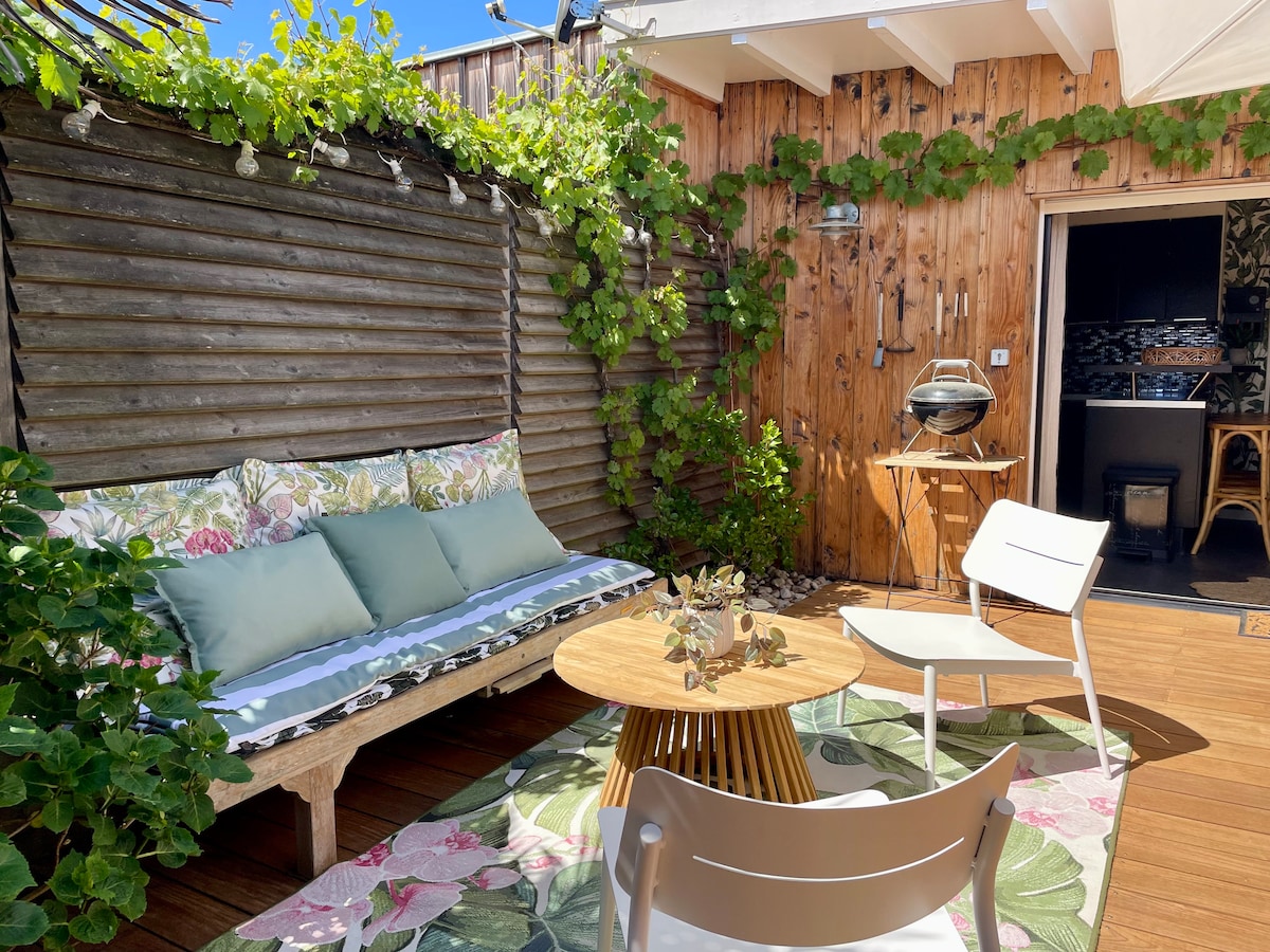 An outdoor patio area features a comfortable seating arrangement with floral cushions on a wooden bench. A round wooden table sits at the center, surrounded by white chairs. Green vines adorn the surrounding wooden walls, complemented by a barbecue grill in the background.