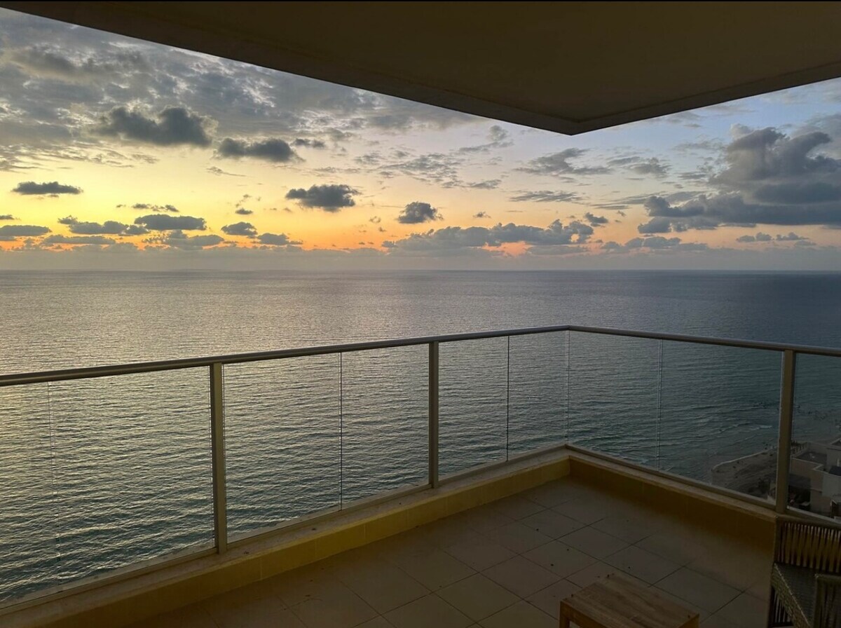 A spacious balcony offers panoramic views of the ocean during sunset. The water reflects hues of orange and blue, while clouds drift across the sky. A small wooden table is positioned on the tiled floor, enhancing the serene outdoor setting.