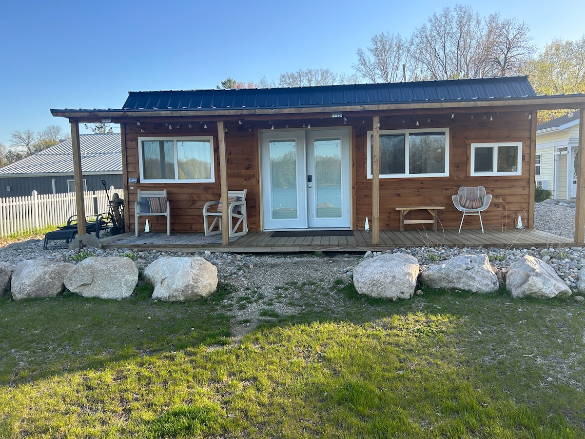 A wooden cabin with a porch is depicted in the image, featuring large glass doors surrounded by two windows. Several comfortable chairs are placed on the porch, complemented by ornamental rocks and a grassy area in front. Trees and a fence are visible in the background.
