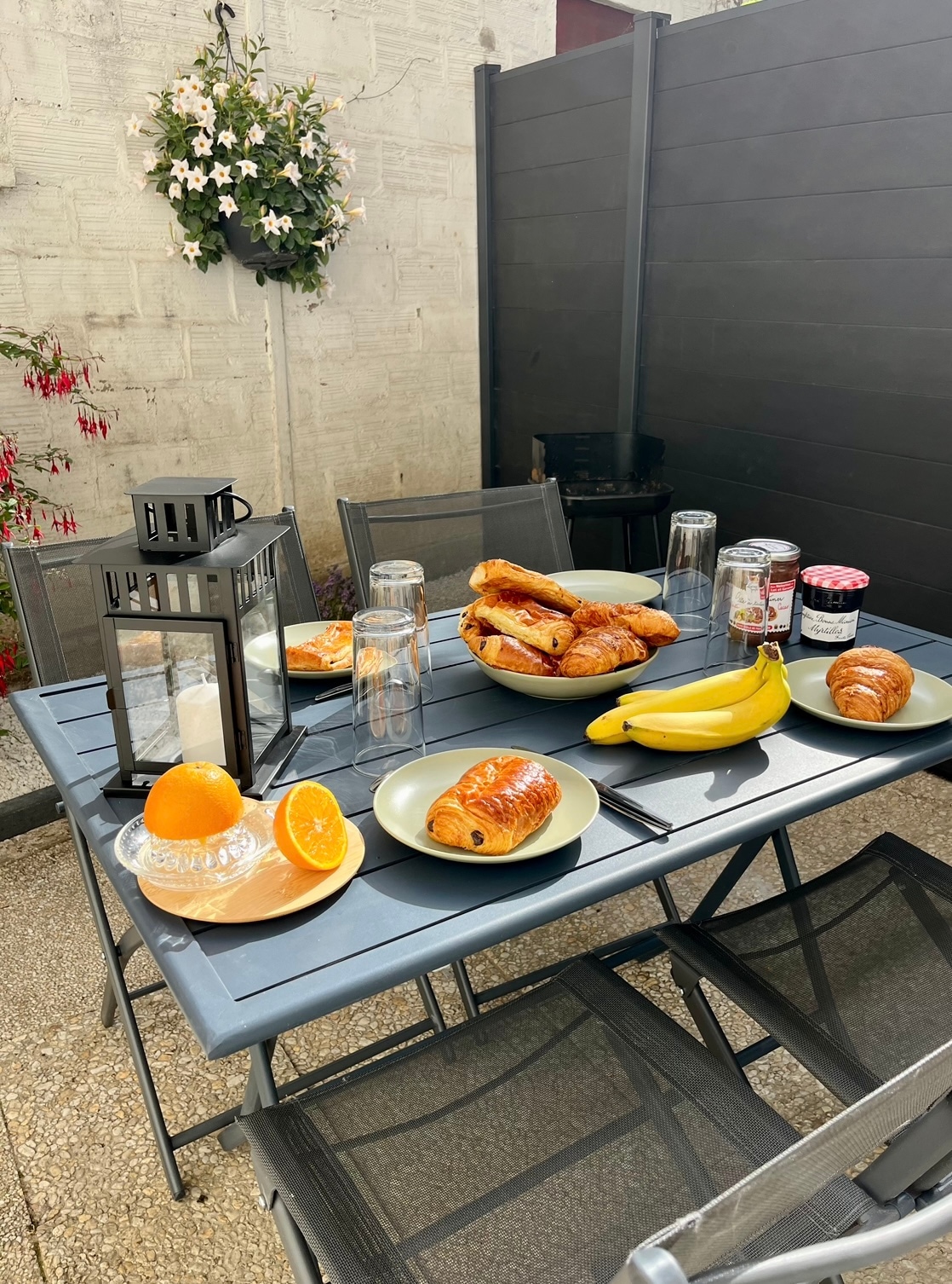 A dining table is set in an outdoor space, featuring plates with assorted pastries and fresh fruit. A lantern is placed on the table, alongside glasses and jars of jam. A flowering plant hangs on the wall in the background, adding a natural touch.