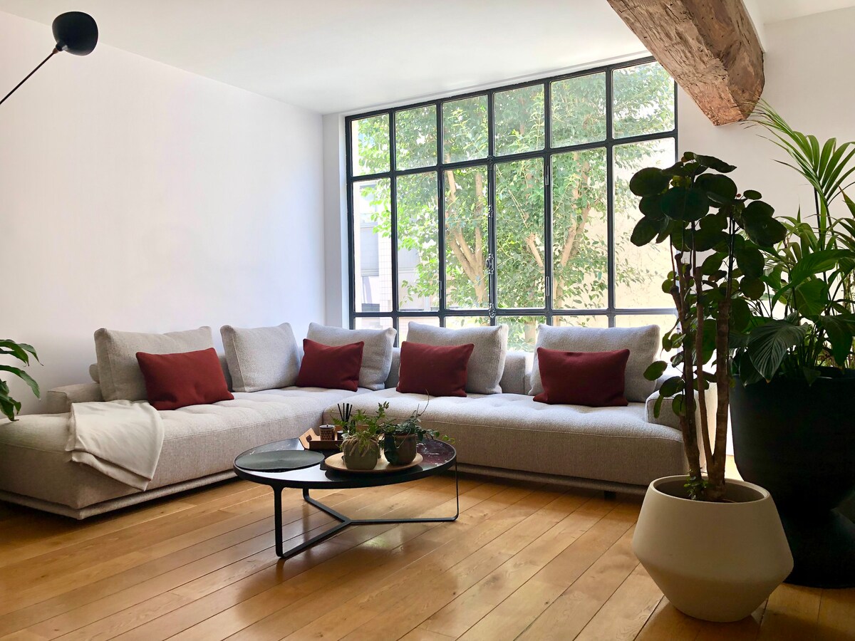A spacious living area is filled with natural light, featuring a large corner sofa with cushions arranged neatly. A minimalist coffee table sits at the center, surrounded by various potted plants that add greenery to the room. Tall windows offer a view of trees outside.