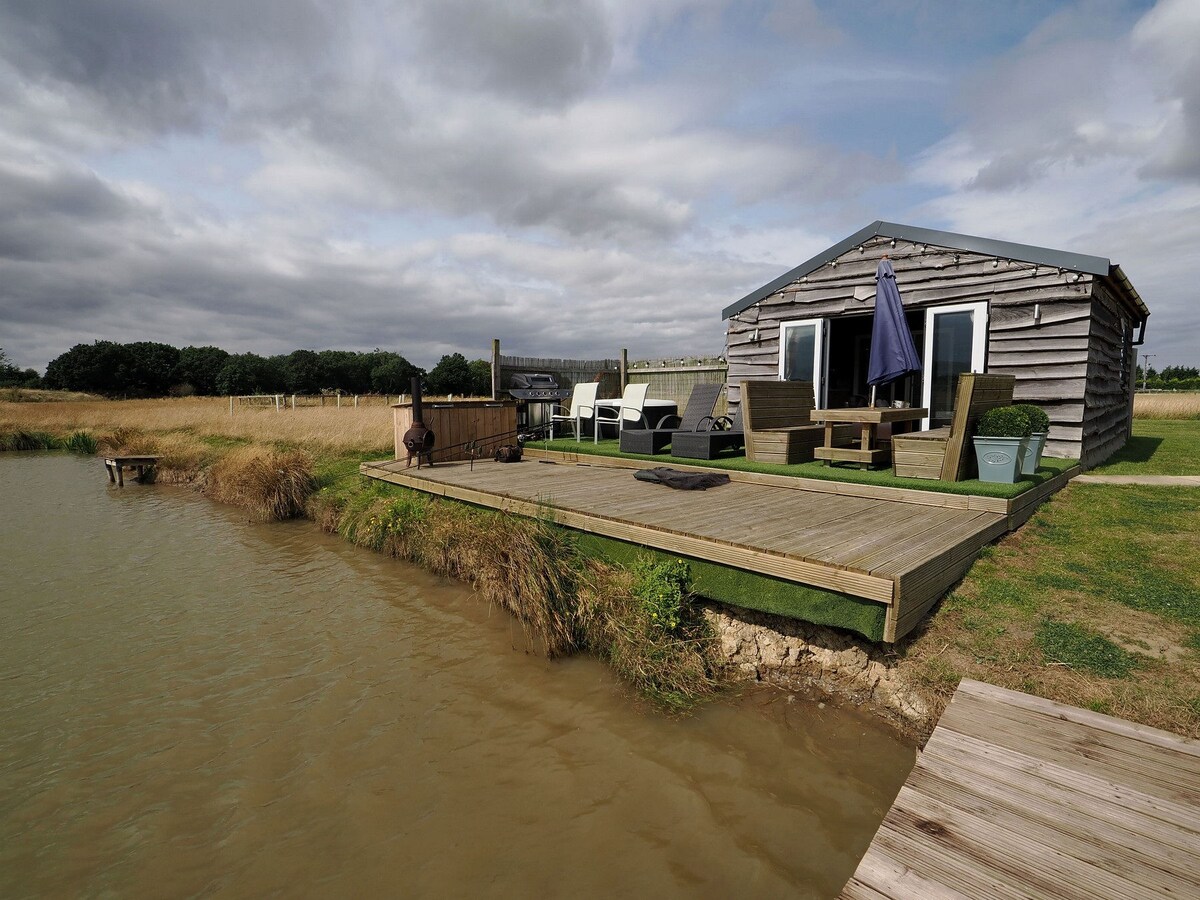 A private cabin is positioned next to a well-stocked fishing lake, surrounded by grassy areas. A spacious wooden deck features outdoor seating and a parasol, providing an inviting space for relaxation. The serene water's edge complements the natural landscape, enhancing the peaceful outdoor setting.