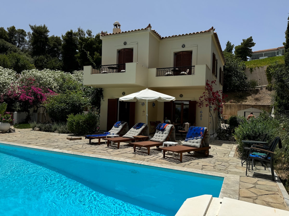 A charming two-story villa is surrounded by lush greenery and flowering shrubs. Sun loungers are positioned near a private pool, alongside a shaded space featuring an umbrella. The balconies are furnished, inviting relaxation with views of the tranquil outdoor area.