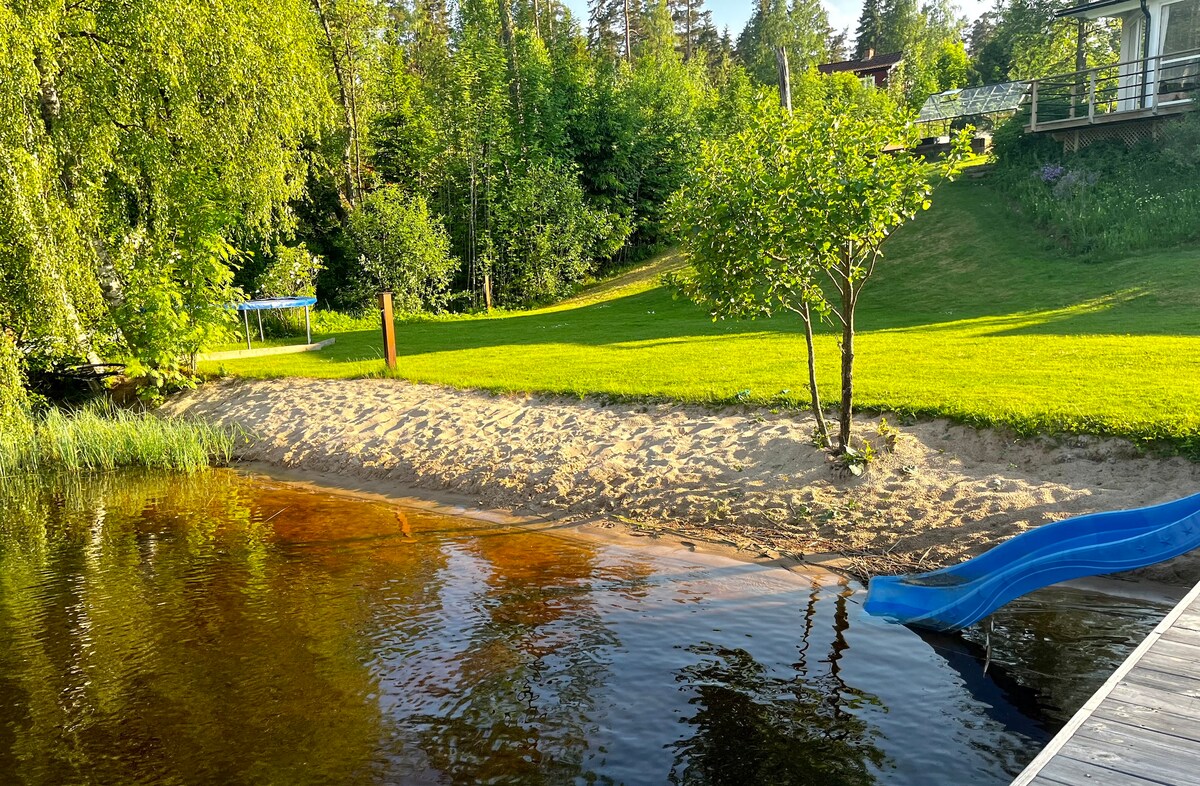 A sandy beach area is framed by lush greenery, leading to a calm lake. A bright blue slide can be seen next to the water, and a grassy slope rises gently behind, with a small structure visible in the background.