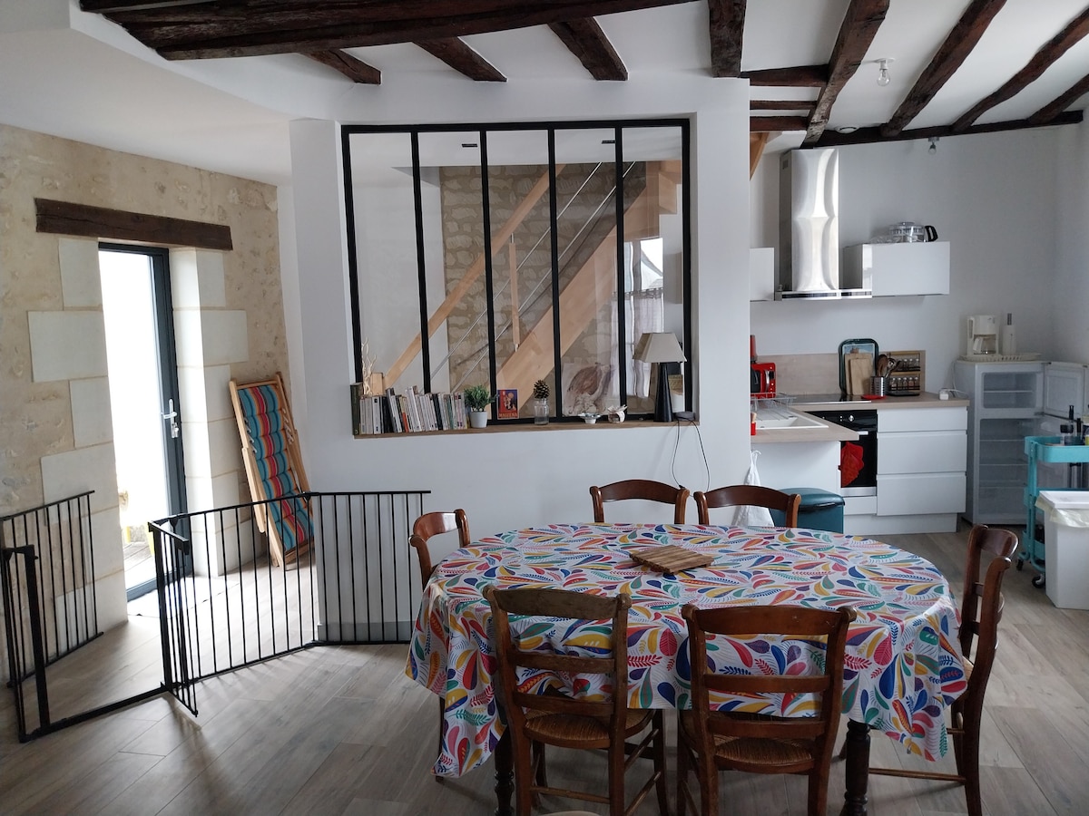 A spacious dining area features a round table adorned with a colorful tablecloth, surrounded by wooden chairs. In the background, an open kitchen with modern appliances is visible. Large windows provide natural light, while a decorative railing enhances the playful charm of the space.