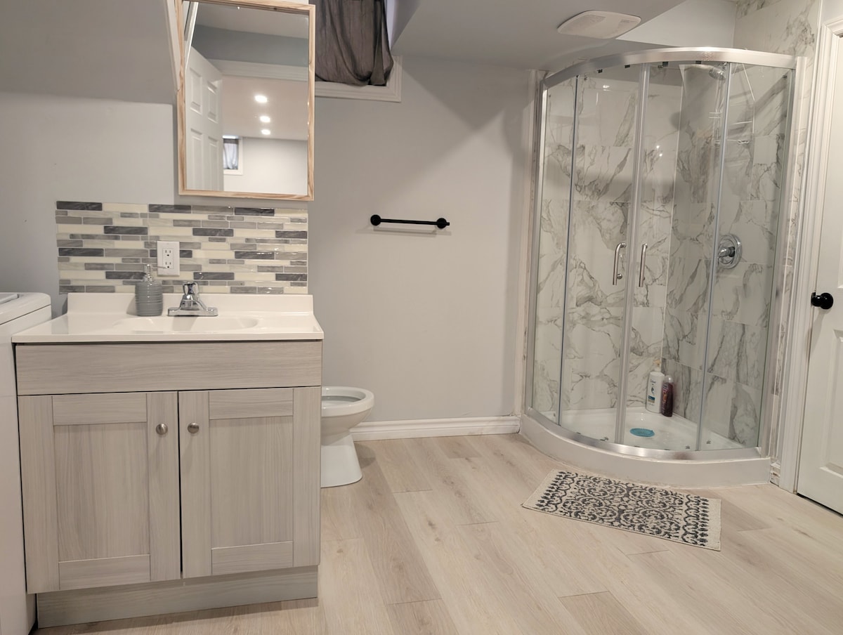 The bathroom features a modern design with a glass shower enclosure and marble-patterned walls. A light-colored vanity with storage sits beside the shower, and a small area rug is placed in front of the shower. Natural light is present through a window above.