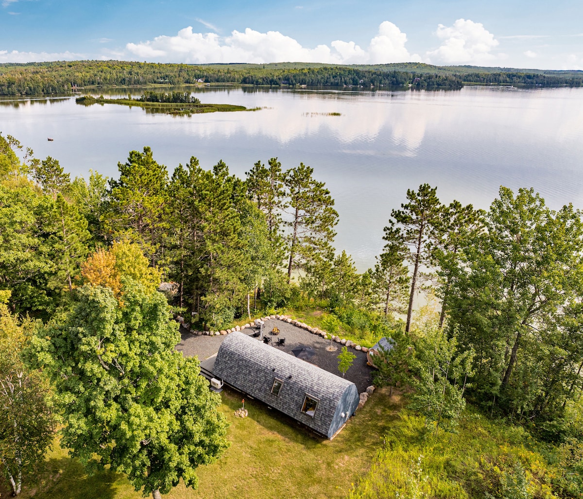 An aerial view of the cabin reveals its unique design and surrounding landscape. The structure sits nestled among trees with a landscaped yard leading to the water's edge. The calm lake reflects the sky, showcasing distant islands and lush greenery.