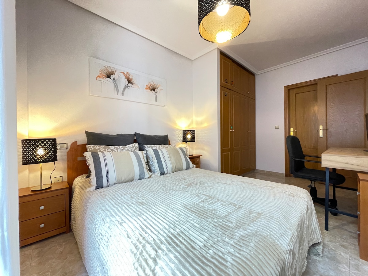 A spacious sleeping area is presented with a large bed covered in a textured blanket. Bedside tables are positioned on each side, featuring decorative lamps. A workspace with a chair is visible, along with a wardrobe and wooden doors in the background, illuminated by soft lighting.