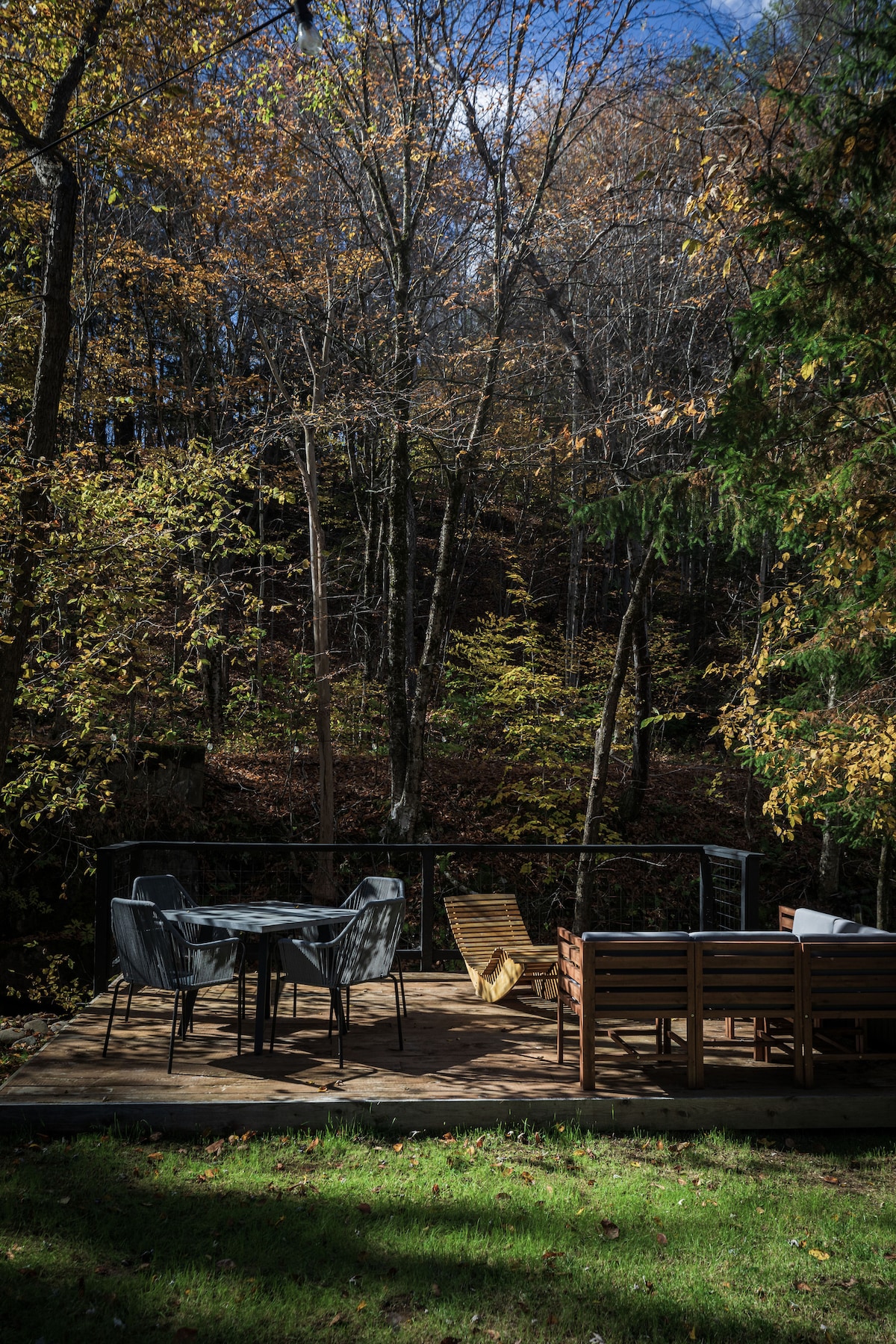 A spacious deck is surrounded by trees showcasing autumn foliage. It features a dining table with four chairs, a lounge chair, and a comfortable outdoor sofa, inviting relaxation amidst nature. The view offers glimpses of the nearby creek and wooded surroundings.