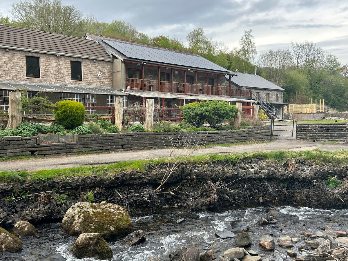 A converted barn is shown, featuring a multi-level design with wooden balconies. The natural landscape surrounds the property, with a small stream in the foreground and a decorative garden area. Trees and greenery create a serene backdrop, enhancing the rustic charm of the setting.