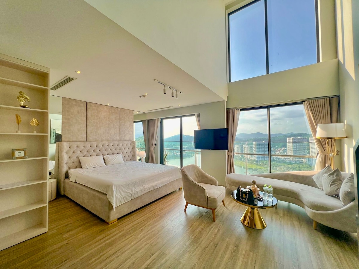 A bright and spacious room features a large bed with a soft headboard, complemented by natural light from floor-to-ceiling windows. A cozy seating area includes a stylish chair and a sleek sofa. A modern entertainment setup is visible on the wall opposite the bed.